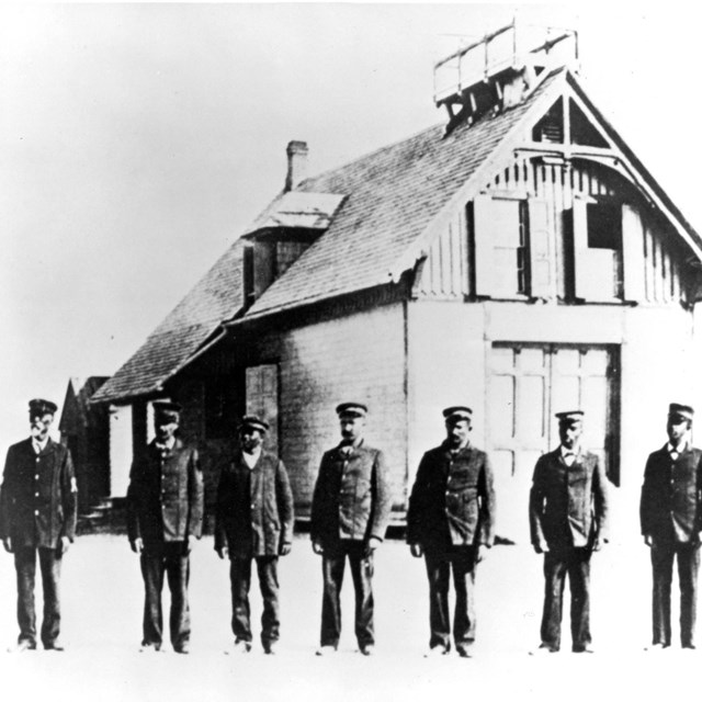 black and white photo of a line of 7 men standing on sand in front of a 2 story wooden building