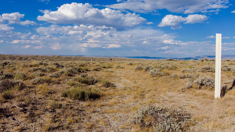 Dirt trail through partially barren sagebrush grasslands with blue skies and white clouds.