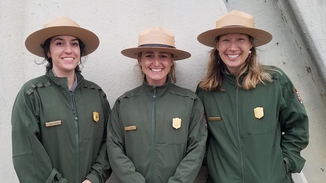 Three uniformed park rangers stand shoulder to shoulder. 