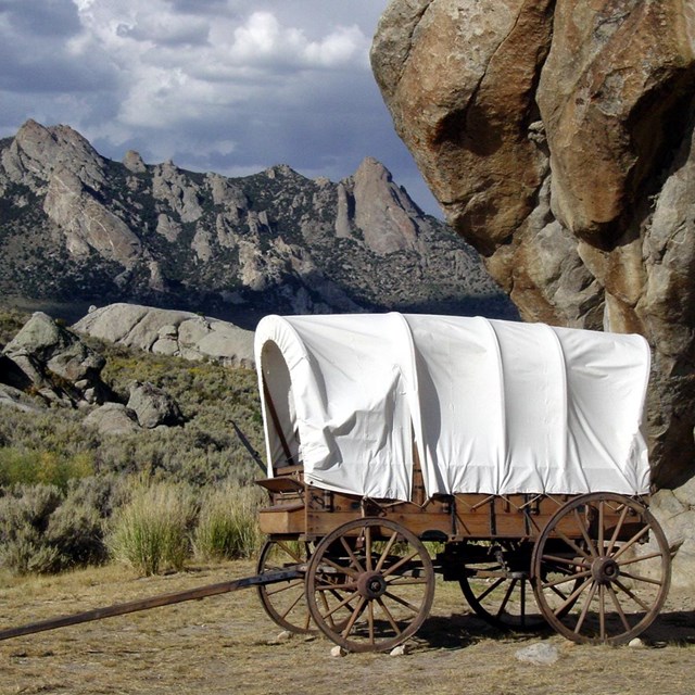 A covered wagon sits in front of a large rock outcropping.