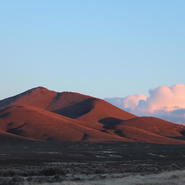 A large, smooth looking hill, colored red from the sunset, with distant billowing clouds.