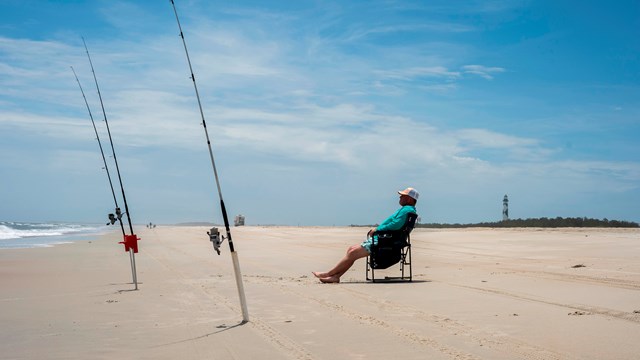 A angler sits on a chair with fishing poles in front of him. 