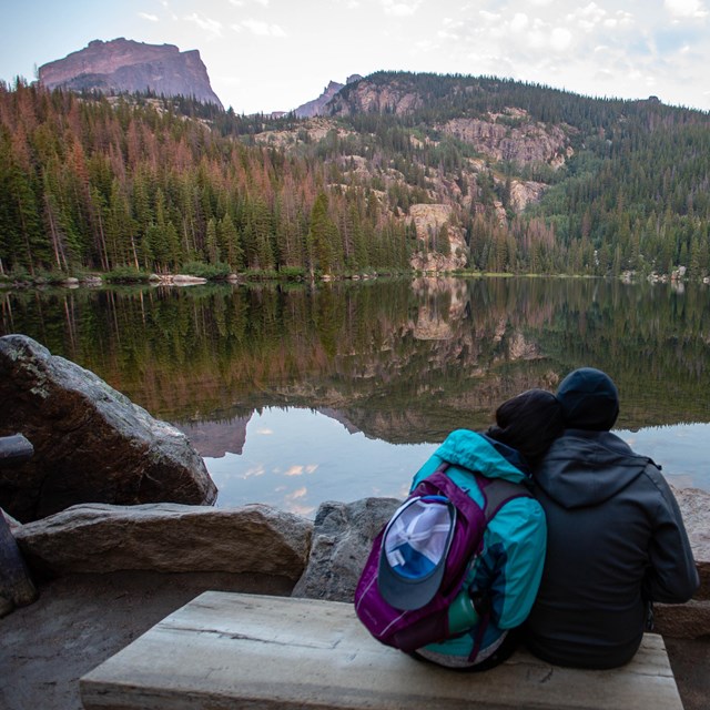 a man and woman sit on a bench looking out onto a lake and mountain during sunrise