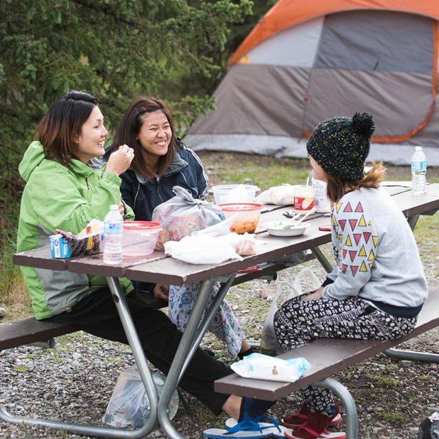 Three women sit at a picnic table eating and laughing. A tent is in the background.