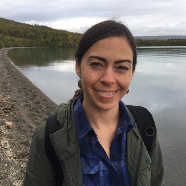 medium shot of a brunette woman smiling at the camera with a shore line and water in the background