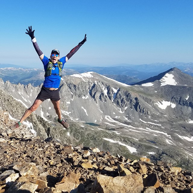 man in running clothes jumps in the air on top of a mountain with snowy peaks in the background