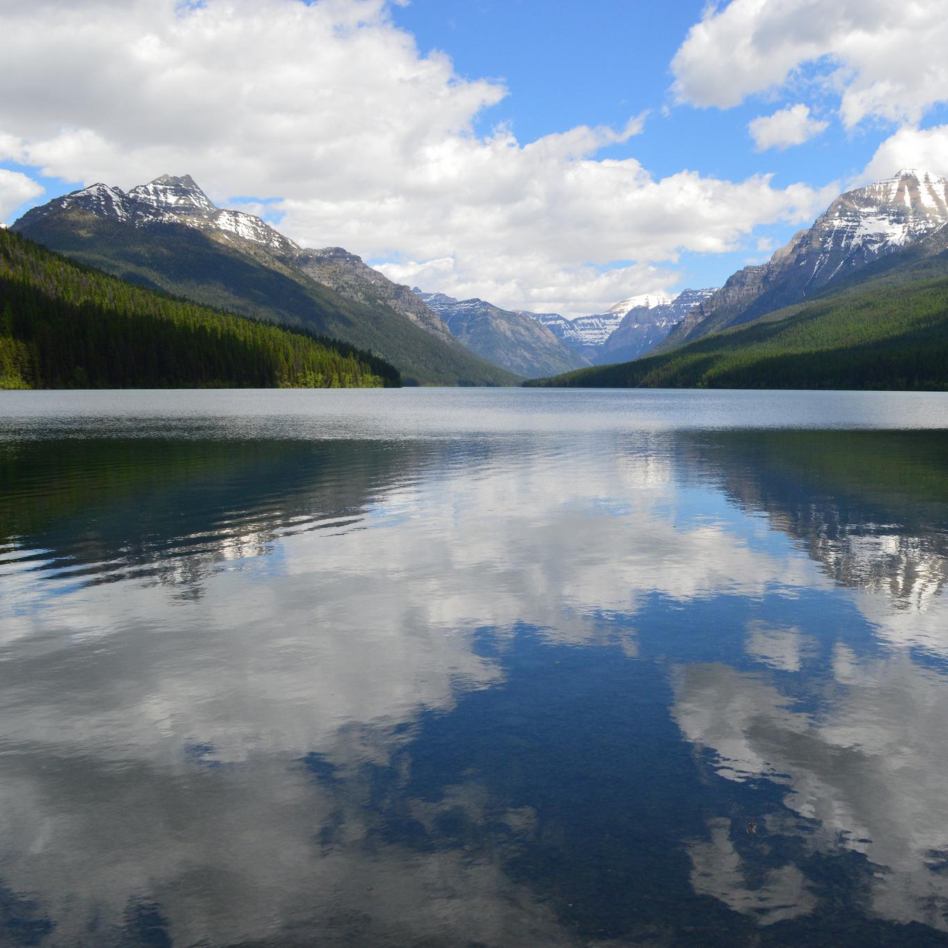 a clear blue lake reflects white clouds with mountains in the background 