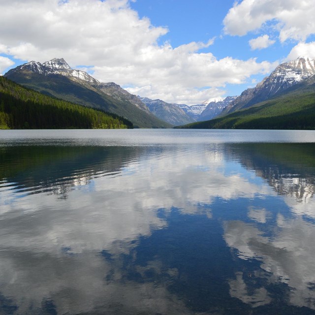 a clear blue lake reflects white clouds with mountains in the background 