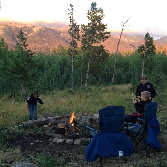3 young kids around a fire pit at dusk with mountains in the background
