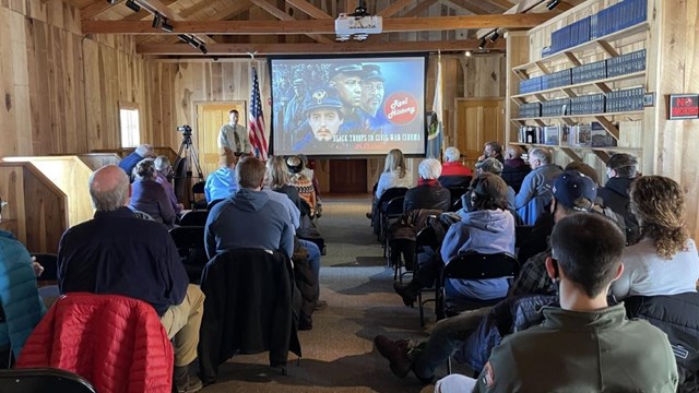 Crowd of seated people watching a speaker and PowerPoint presentation