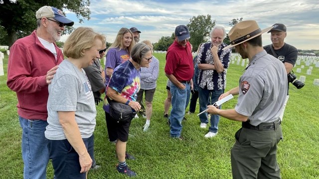 Ranger showing an image to crowd of visitors in green cemetery field.