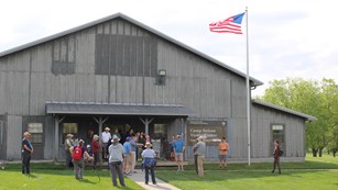 A large group of people stand in front of a wooden building with a US flag flying.