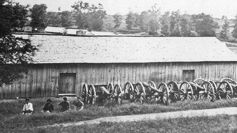 Four soldier sit beside a row of cannons outside a wooden ordnance warehouse
