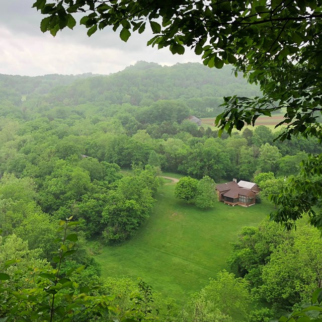 Looking over a large building and field surrounded by lush forest and rolling hills