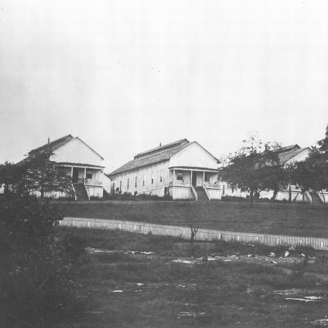 Rectangular wooden buildings with trees and a field in front during the Civil War.