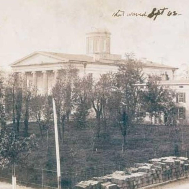 A large state house building with a grassy field, trees, and fence in front during Civil War.