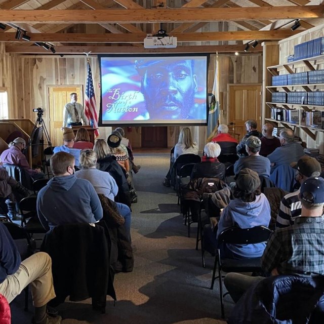 Rows of seated people indoors watching a speaker and projector image.