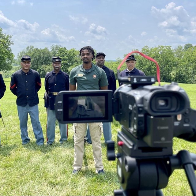 Video camera in front with line of men, mostly in Civil War uniforms, in background.