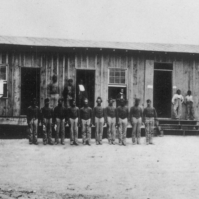 African American soldiers standing in front of wooden building with windows during the Civil War.