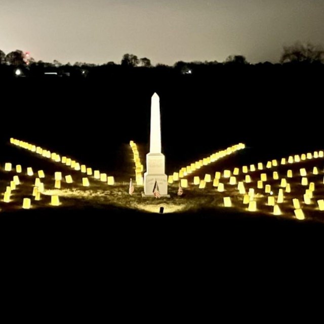 Stone obelisk with lines of luminaria stretching away from it at night.