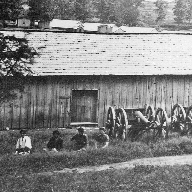 Four soldier sit beside a row of cannons outside a wooden ordnance warehouse