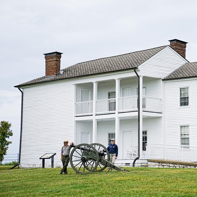 A National Park Service Ranger and an intern stand in the front yard of a historic house. 