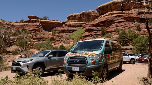 Two cars parked at the Elephant hill trailhead, sandstone formations tower above them