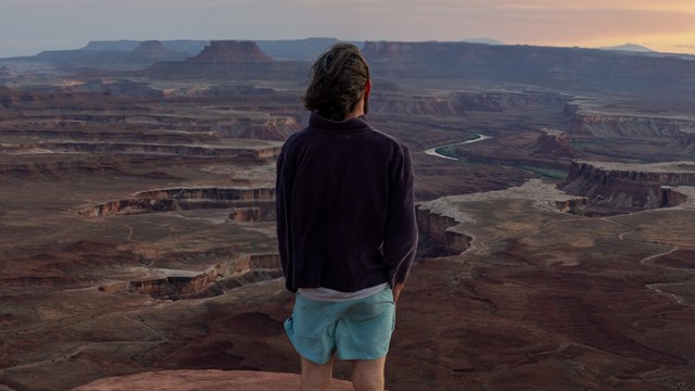 A person stands at the edge of a canyon, overlooking layers of canyons at sunset. 