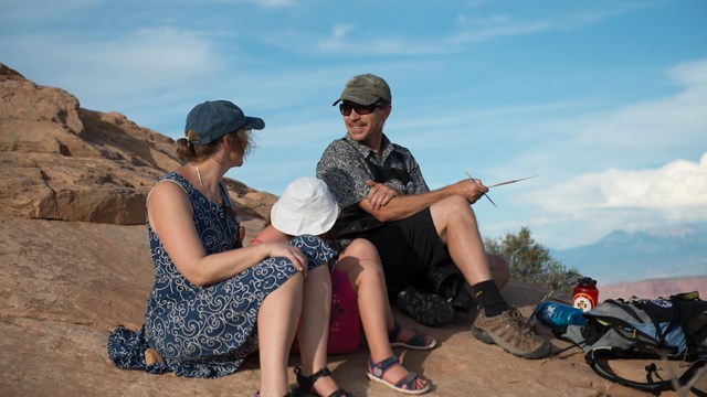 Two adults and a child site on a rock looking at artwork.