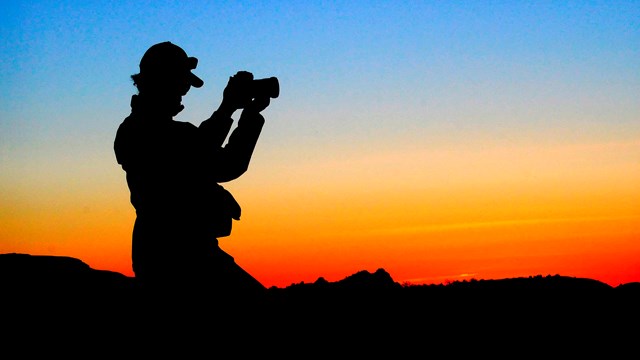 A silhouette of a person holding a camera against a blue, yellow, and orange sky 