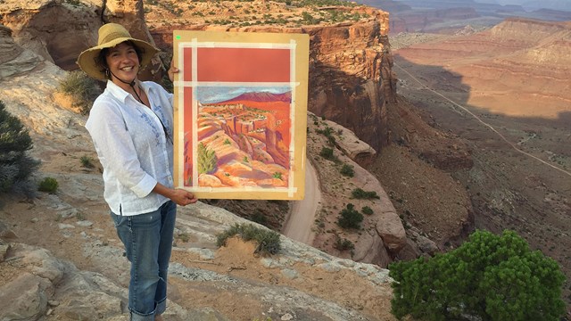 a woman at a scenic overlook holds up a painting