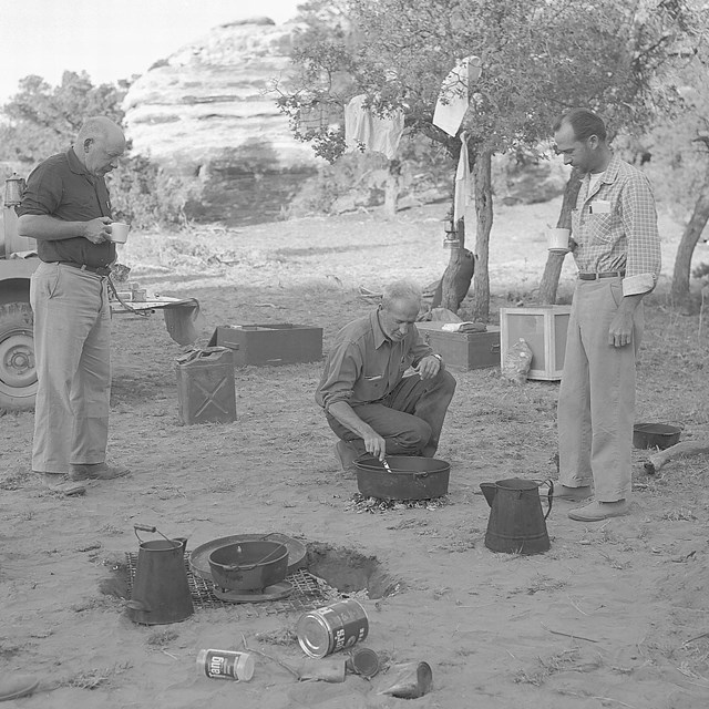 Bates Wilson kneeling over a campfire stirring a pot. 3 Men watch