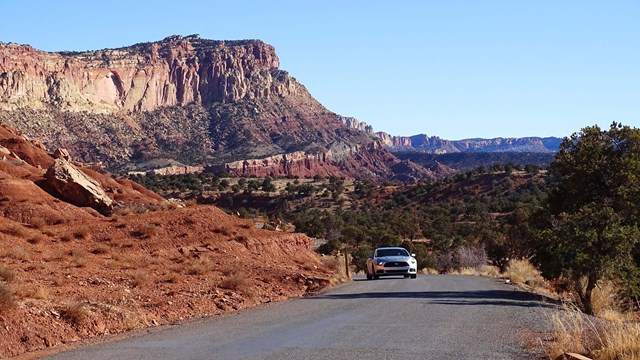 A car drives a narrow road with tall red cliffs rising on one side of the road.