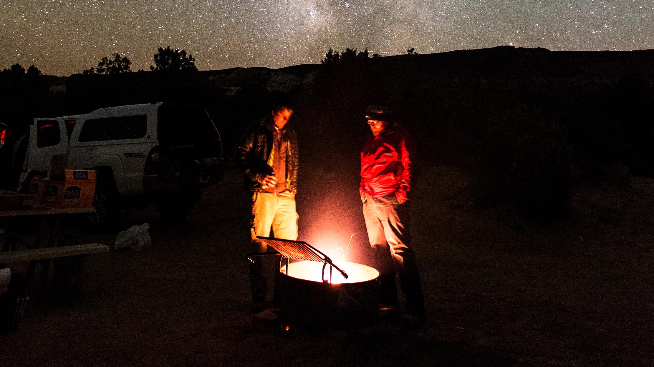 Two people stand beside a campfire, with the Milky Way above them. 