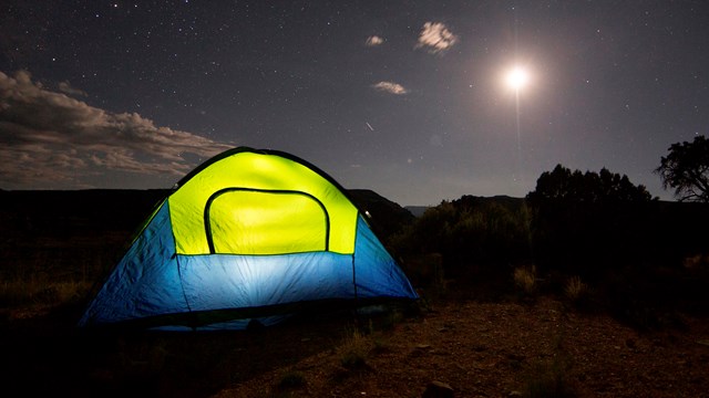 Green and blue tent with a light inside set up at night, with dark skies and a full moon above.