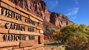 Red stone wall with the words "Visitor Center Capitol Reef National Park". Cliffs and trees behind