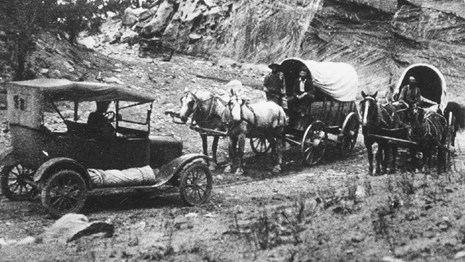 Black and white photo of old fashioned car meeting two wagons pulled by horses in a narrow canyon.