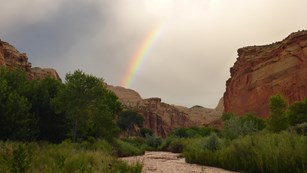 Reddish brown river flowing between green banks, with cliffs, a stormy sky, and rainbow.