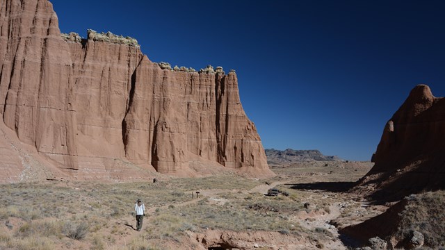 Person hiking on dirt trail below big red cliffs and blue sky, with a car parked in the distance. 