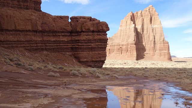 Large red, triangular monolith, reflected in a puddle, with blue sky above, and red rock to the side