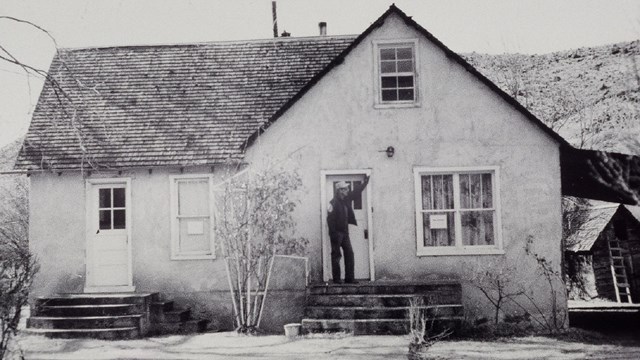 Black and white photo of a man standing on the porch of a house. 