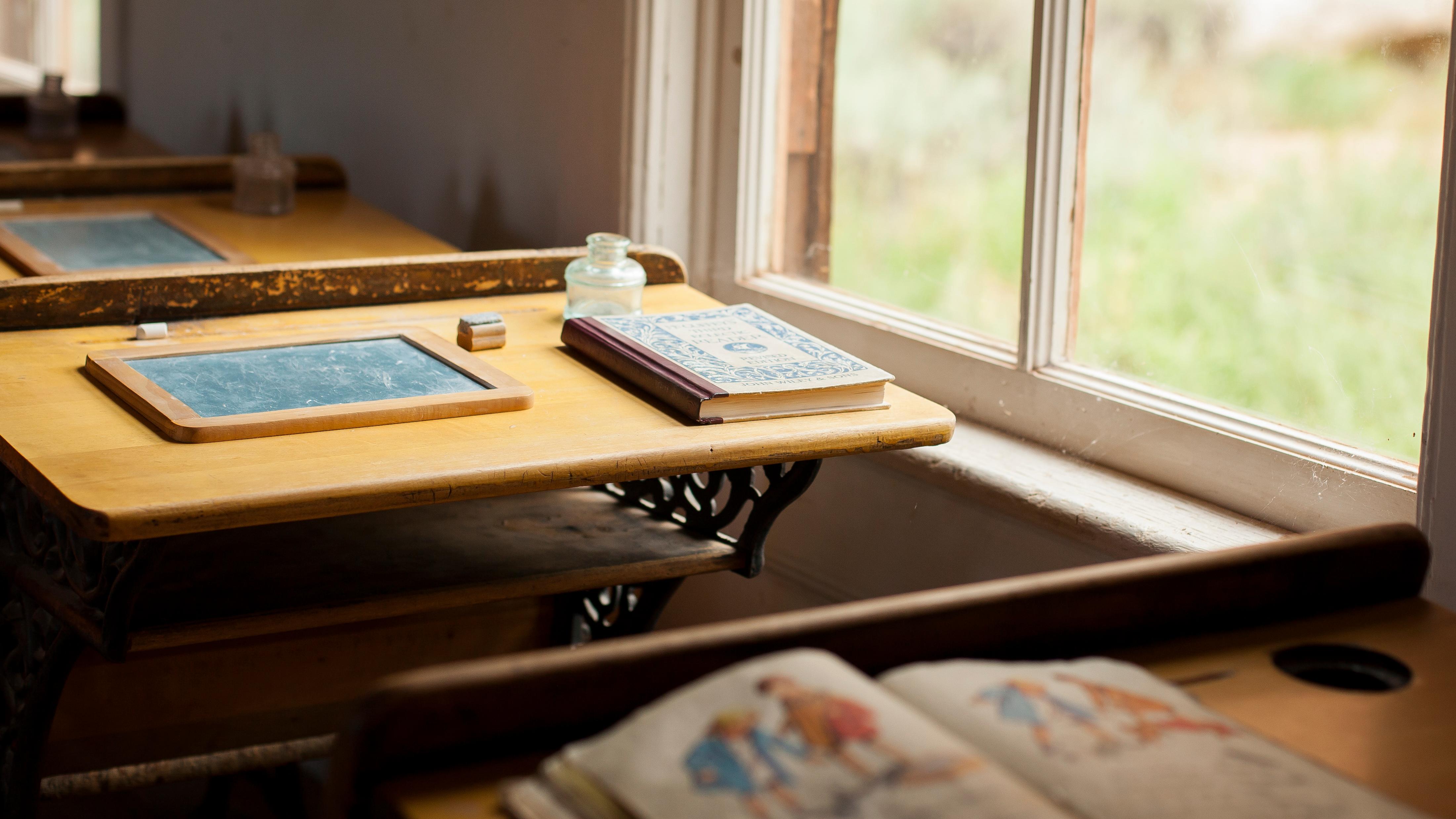 Photo of old-fashioned school books on old-fashioned desks beside windows.
