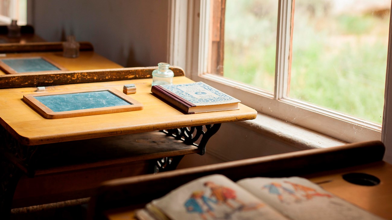 Photo of old-fashioned school books on old-fashioned desks beside windows.