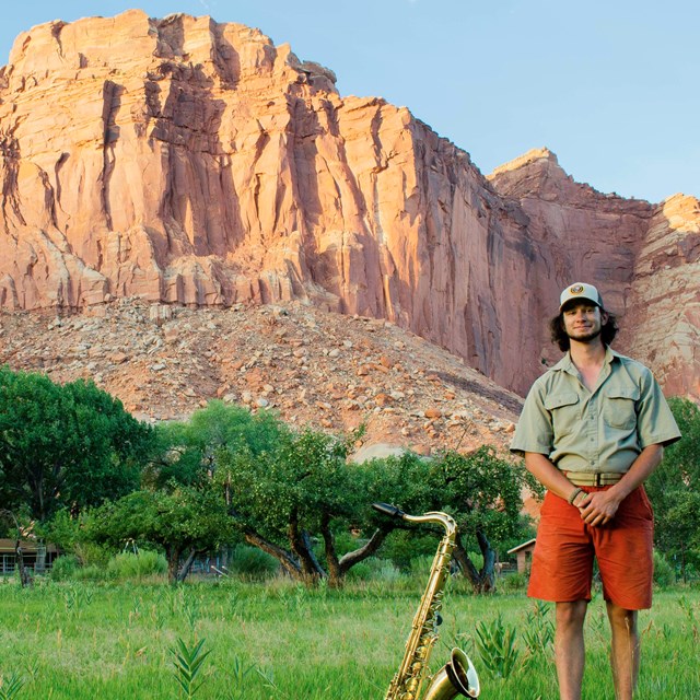 A man standing in a green field next to a gold saxophone with orange rock cliffs in background.