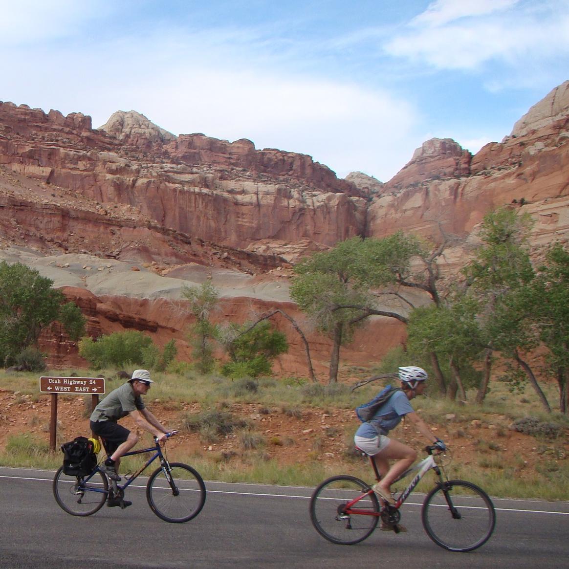Two bike riders on paved highway, with scenic rock formations and blue sky in the background. 