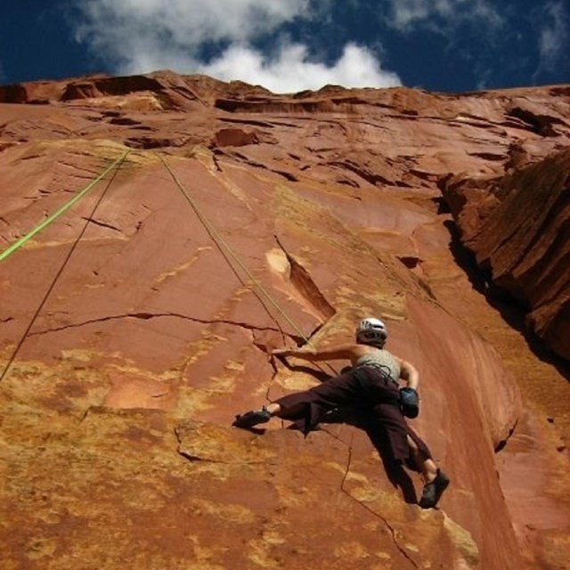 Person climbing red cliff wall with blue sky and clouds above.