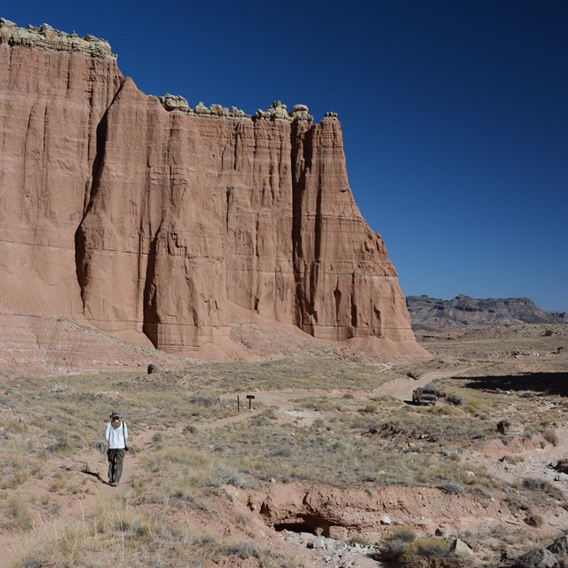 Person hiking on dirt trail below big red cliffs and blue sky, with a car parked in the distance. 