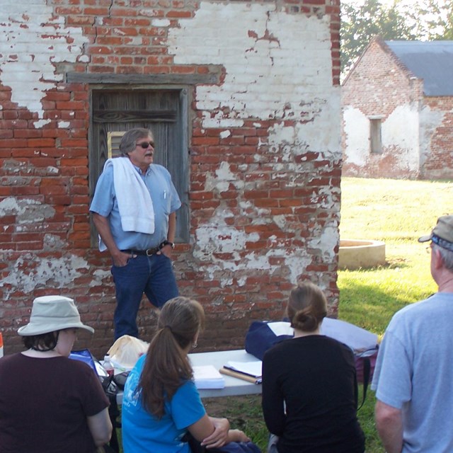 Adult learners gathered around table, listening to speaker in front of historic cabin.