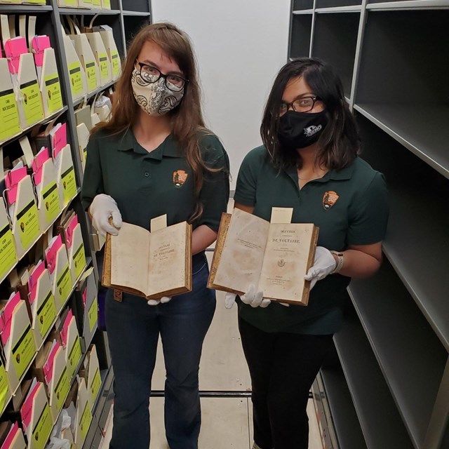 Interns stand with open books, next to shelf of boxes of artifacts. 