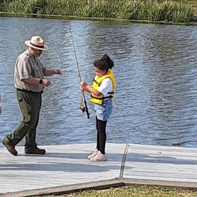 Ranger helps child with fishing pole on river. 
